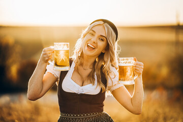 Wall Mural - Pretty happy blonde in dirndl, traditional festival dress, holding two mugs of beer outdoors in the field