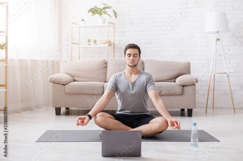 Meditation and yoga. Guy with closed eyes sits in lotus position on mat, with laptop