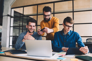 Millennial hipster guys collaboration together in coworking space sitting at desktop with laptop while one of them making photos of computer screen, man reading sms message via digital tablet