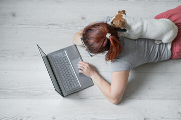 A European red-haired woman is lying on the floor with her dog and is typing on a laptop. Female student studying remotely at home in quarantine. A loyal puppy Jack Russell Terrier. Exam preparation.