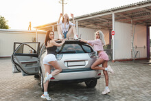 Three Hot Girls In Frank Outfits Pose On The Car In The Rays Of The Sunset. Women Pose At The Car Wash. Professional Models Next To Expensive Cars.