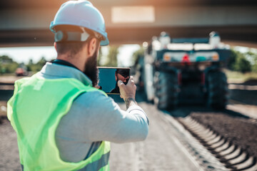 Wall Mural - Young male road construction worker on his job. He uses smart phone or tablet to make video log about work.Bright sunny day. Strong light.