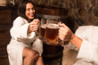 happy young caucasian women in bathrobe sit next to pool at sauna, drinking cold beer. 