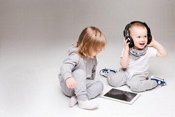 Stylish brothers and sisters two year old sitting on floor at gray studio, listening music at headphones, having fun together. Little clever boy and girl with tablet, spending time and playing.