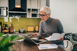 © bnenin - Portrait of a senior woman working with laptop and documents in the kitchen.