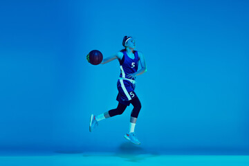 Wall Mural - In high jump. Young caucasian female basketball player training, prcticing with ball isolated on blue background in neon light. Concept of sport, movement, energy and dynamic, healthy lifestyle.