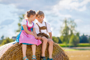 Wall Mural - Two kids in traditional Bavarian costumes in wheat field. German children sitting on hay bale during Oktoberfest. Boy and girl play at hay bales during summer harvest time in Germany. Best friends.