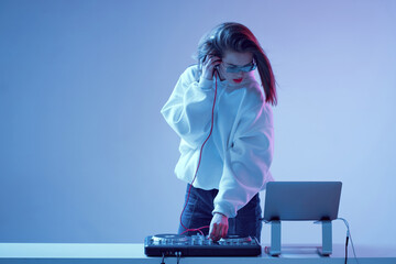 Cool young girl DJ mixes music on a mixing console and laptop, in stylish clothes, glasses on a neon background.