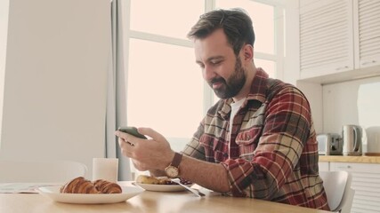 Wall Mural - Handsome young positive bearded man at the kitchen indoors at home having a breakfast while using cellphone and drinking tea or coffee