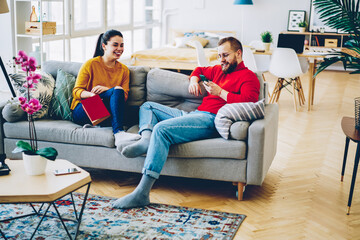 Wall Mural - Cheerful couple in love having fun communicating with each other at comfortable home interior,happy hipster guy joking with his girlfriend reading message from smartphone, family spending time at home