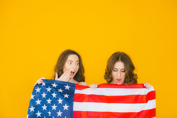 American girls. Two playful young women gesturing peace sign and holding American flag while standing on yellow background