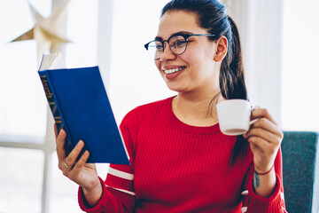 Wall Mural - Cheerful brunette woman in spectacles for vision correction enjoying free time on reading interesting novel, smiling hipster girl holding coffee cup and book satisfied with free time at home .