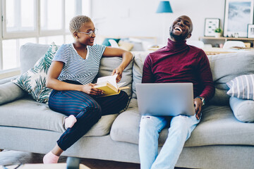 Wall Mural - Cheerful african american hipster guy laughing during conversation with girlfriend at home interior, couple sitting at comfortable sofa on leisure man using netbook while his girlfriend read book