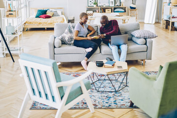 Wall Mural - Romantic african american couple discussing new book spending free time together at home interior, dark skinned family talking to each other about literature using netbook 