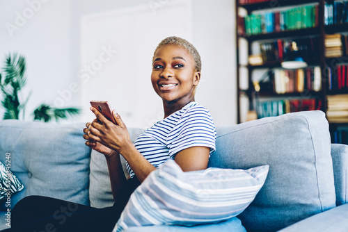 Fotografía Cheerful african american woman satisfied with wireless internet at home connecting smartphone for chatting and blogging,happy dark skinned hipster girl using mobile phone for sending message