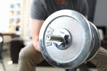 Close-up of male person lifting heavy metal dumbbell sitting on chair. Strong fit body. Training and exercise for best result. Home gym and active lifestyle concept