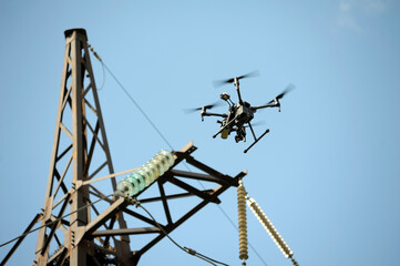 Pro quadcopter with camera aboard flying in a sky, electric pylon, cables and insulators on the background