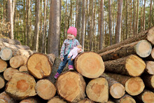 Smiling Little Girl With Doll Sitting On Stack Of Wood In The Forest