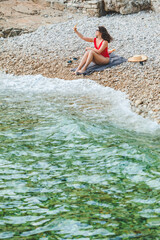 Wall Mural - young pretty woman in red swimsuit sitting at rocky beach with phone taking selfie picture