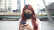 © S photographer - A woman dyed in red hair wearing a mask and standing playing with a mobile phone in the city. 4k Resolution.