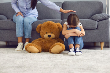 upset little girl crying on floor near teddy bear while mother comforting her at home. parent soothi