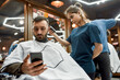 © Svitlana - Businessman visiting barbershop. Young handsome bearded guy sitting in armchair in the barbershop and using his mobile phone while a professional barber girl doing haircut for him