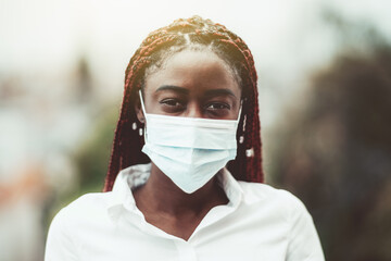 An outdoor portrait of a young African female with chestnut braids and in a virus protective mask on her face; masked black woman outdoors with protection against influenza and pandemic threat