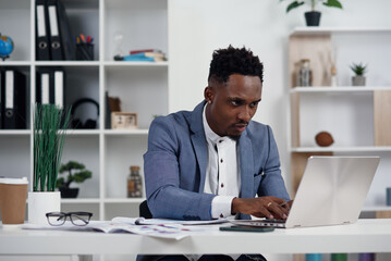 Wall Mural - Businessman with laptop. Young African businessman is typing something on laptop in his office.