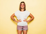 Fototapeta Panele - Young beautiful woman looking at camera.Trendy girl in casual summer white T-shirt and jeans shorts in round sunglasses. Positive female shows facial emotions. Funny model isolated on yellow