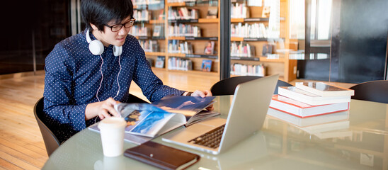 Wall Mural - Young Asian man university student reading book while working with laptop computer in library. Self learning and education research. Scholarship for educational opportunity concepts