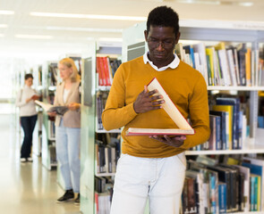 Wall Mural - African-american man reading book while standing near bookshelves in public library