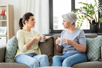 Wall Mural - family, generation and people concept - senior mother with adult daughter drinking coffee or tea and talking at home