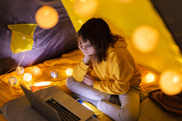 Wall Mural - young girl student using laptop for homeschooling in a home made tent with glowing light balls.