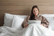 © lenblr - beautiful girl in bathrobe studies online with tablet sitting in soft bed in hotel room during self-isolation