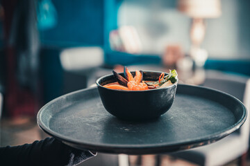 Poster - pan asian food. waiter hands holding Tom Yam soup in black bowl