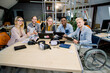 © sofiko14 - Creative team of diverse business people sitting at office table during meeting, looking at camera with smile, passionate about work. A handicapped man in a wheelchair, chief with his team