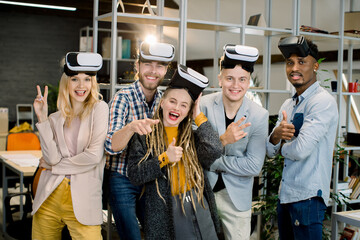 Wall Mural - Five cheerful multiethnic friends coworkers enjoying their work with VR goggles at modern creative office room. Young people posing with vr headset smiling and gesturing to camera