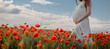 Close up a pregnant woman in white dress holding with her hands her belly in a red poppies flowers field