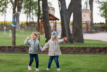 Cute little children enjoy a warm summer day in the fresh air: they run around the green lawn of a public park and try to catch soap bubbles