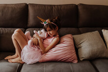 Side View Of Barefoot Girl In Unicorn Costume Embracing Plush Toy While Resting On Comfortable Couch At Home
