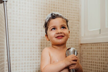 Smiling Little Child With Foam On Head Standing In Bathroom With Shower And Singing While Looking Away