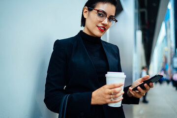 Half length portrait of positive female proud ceo in formal wear holding takeaway cup with hot caffeine beverage and smartphone gadget and looking at camera while waiting partner on urban setting