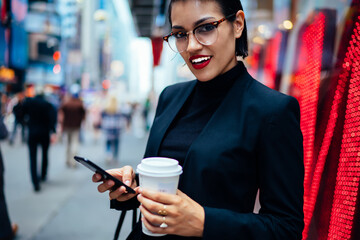 Half-length portrait of prosperous businesswoman satisfied with career and occupation standing outdoors checking mail on mobile, confident female manager in eyeglasses looking at camera sending sms.