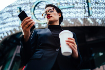 Wall Mural - Serious female entrepreneur with red lips updating application on mobile while strolling on street, confident businesswoman in formal wear making money transaction online on coffee break outdoors.