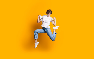 Jumping caucasian woman in jeans and white t-shirt smiling and advertising something on a yellow studio wall