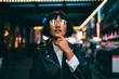 Young trendy dressed hipster in eyewear with night city light reflection fascinated with beautiful illumination, gorgeous woman in leather jacket wondering while standing