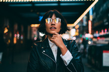 Young Trendy Dressed Hipster In Eyewear With Night City Light Reflection Fascinated With Beautiful Illumination, Gorgeous Woman In Leather Jacket Wondering While Standing