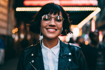 Wall Mural - Half-length portrait of cheerful hipster girl spending time on night street of New York listening music in earphones, gorgeous young woman looking at camera enjoying favorite playlist at evening city