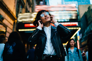 Positive hipster girl in trendy clothes listening favorite song from playlist while strolling on crowded city street in evening, young woman enjoying sound in earphones walking at night downtown