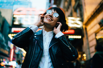 Wall Mural - Smiling young woman in gorgeous apparel dancing while listening favorite song in earphones walking on night city street, happy hipster girl enjoying music walking on Manhattan with illumination lights